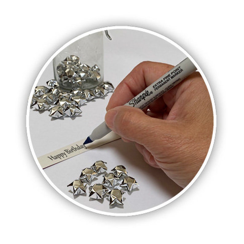 Puede incluir: Una mano escribe "Feliz cumplea&ntilde;os" en una tarjeta blanca con un marcador Sharpie plateado. Estrellas de origami plateadas est&aacute;n esparcidas alrededor de la tarjeta.