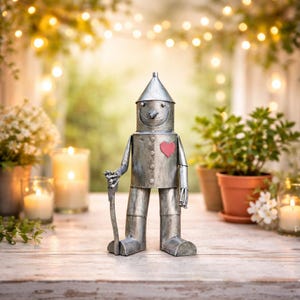 Może przedstawiać: Metalowa rzeźba Blaszaneho Drwala z Czarnoksiężnika z Oz, z stożkowym kapeluszem, czerwonym sercem i laską. Srebrna rzeźba stoi na drewnianej powierzchni. Tło obejmuje rośliny, świece i łańcuchy świetlne.