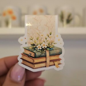 Könnte beinhalten: Ein Aufkleber mit einem Stapel Bücher, der mit einem Blumenarrangement verziert ist. Die Bücher haben eine Vintage-Ästhetik mit goldfarbenen Seiten und einem grünen Einband. Ein beiges Band umschließt die Bücher. Der Hintergrund ist hellbeige mit einem floralen Muster.