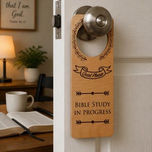 May include: A light-colored wooden door hanger featuring a circular cutout at the top. The design includes a laurel wreath, a banner with the text "Quiet Please," and the words "BIBLE STUDY IN PROGRESS" centered between two decorative arrow motifs.