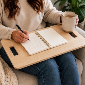 May include: A rectangular light-toned wooden lap desk featuring rounded corners and two integrated cutout handles on the sides. The flat surface supports an open spiral-bound notebook with lined pages and a ceramic mug filled with a light brown beverage.