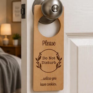 May include: A light-colored wooden door hanger with a circular cutout for a doorknob. The text reads: Please, Do Not Disturb, within a leaf wreath design, and below it, the text: ...unless you have cookies. The sign is hanging on a silver doorknob.