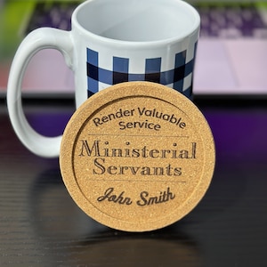May include: A white ceramic mug with a blue and white plaid pattern, next to a round cork coaster. The coaster is engraved with the words "Render Valuable Service," "Ministerial Servants," and "John Smith."