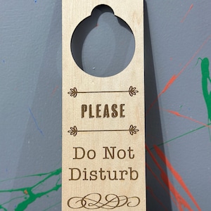 May include: A wooden door hanger with a hole at the top for hanging on a door knob. The hanger is light brown and has the text "PLEASE DO NOT DISTURB" engraved on it.