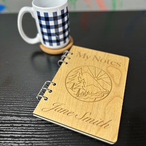 May include: A wooden spiral bound notebook with the words "My Notes" engraved on the cover. The cover also features a laser-engraved design of a mountain scene with trees and a sun. The name "Jane Smith" is engraved below the design.