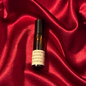May include: A small, brown glass roll-on bottle with a black cap, set against a vibrant red satin background. The bottle has a white label with the text "Mantecadito" and ingredient information. The overall aesthetic is simple and elegant.