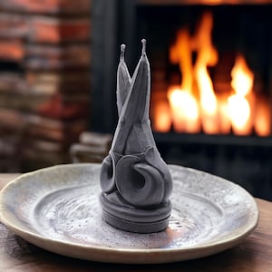 May include: A dark gray candle shaped like a pair of scissors, standing on a ceramic plate with a gold rim. The candle has two wicks and is set against a blurred background of a fireplace with a burning fire.
