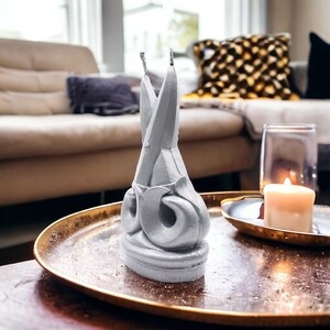 May include: A silver-colored candle shaped like a pair of scissors. The candle has two wicks and sits on a gold-colored tray. A lit candle in a glass holder and a decorative pillow are in the background.