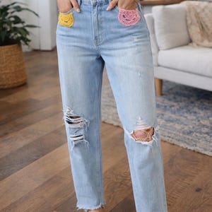 Op de afbeelding: Lichtblauwe denim jeans met distressed details bij de knieën en rafelige zomen. De jeans hebben gehaakte versieringen op de voorzakken, één gele en één roze. De persoon draagt bruine sandalen.