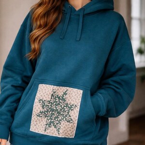 May include: Teal blue hoodie with a kangaroo pocket and drawstring hood. The pocket features a star-shaped design made of green leaves on a beige background. The hoodie is made of a soft material.