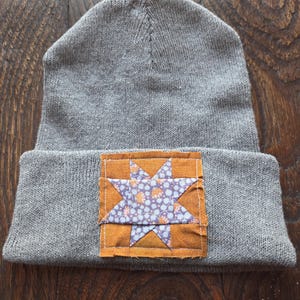 May include: Gray knit beanie with a folded brim. A square patch featuring a star design is sewn onto the front. The star is made of purple and orange patterned fabric, set against an orange background.
