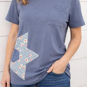 Può includere: T-shirt blu con maniche corte e taschino sul petto. La maglietta presenta una toppa decorativa a forma di stella sul davanti, realizzata in tessuto fantasia con disegni floreali blu, bianchi e rossi. La persona indossa jeans blu.