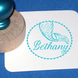 May include: A turquoise blue rubber stamp with a wooden handle. The stamp features a mermaid tail design with the name "Bethany" in a decorative script font.