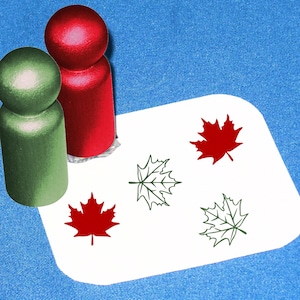 May include: A white card with four maple leaf stamps. Two of the stamps are red and two are green. There are two red wooden stamps, one green wooden stamp, and a white card on a blue surface.
