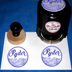 Puede incluir: Un sello de goma autoentintado con mango de madera. El sello presenta un dise&ntilde;o azul y blanco de olas y el nombre "Ryder" en letra cursiva. El sello est&aacute; hecho por "Remarkable Marks".