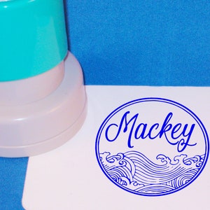 Puede incluir: Un sello autoentintado azul y blanco con el nombre "Mackey" en letra cursiva. El sello presenta un dise&ntilde;o de olas estilizadas.