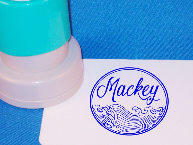 Puede incluir: Un sello autoentintado azul y blanco con el nombre "Mackey" en letra cursiva. El sello presenta un dise&ntilde;o de olas estilizadas.
