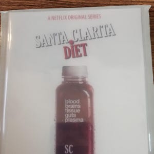 Könnte beinhalten: Eine transparente Plastikhülle mit einem Werbebild für die Netflix-Serie "Santa Clarita Diet". Das Bild zeigt eine Flasche mit roter Flüssigkeit, die mit "blood, brains, tissue, guts, plasma" beschriftet ist. Der Text "Flesh squeezed" steht unten.