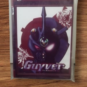 Puede incluir: Una tarjeta coleccionable de Anime Collection, con un personaje Guyver morado y negro. La tarjeta está en una funda de plástico transparente. Se ve el título "Guyver" y el texto "The Bioboosted Armor".