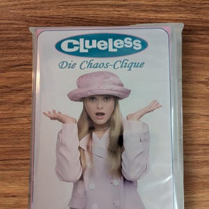 Może przedstawiać: Zapakowany przedmiot z tytułem filmu "Clueless" i tekstem "Die Chaos-Clique". Obraz przedstawia osobę w różowym kapeluszu i płaszczu z wyciągniętymi ramionami. Tło to drewniana powierzchnia.