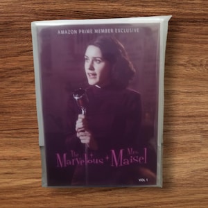 Puede incluir: Una funda de DVD de "The Marvelous Mrs. Maisel Vol. 1" en una funda de plástico transparente. La portada presenta a una mujer sosteniendo un micrófono, con el texto "Amazon Prime Member Exclusive". El fondo es morado oscuro.
