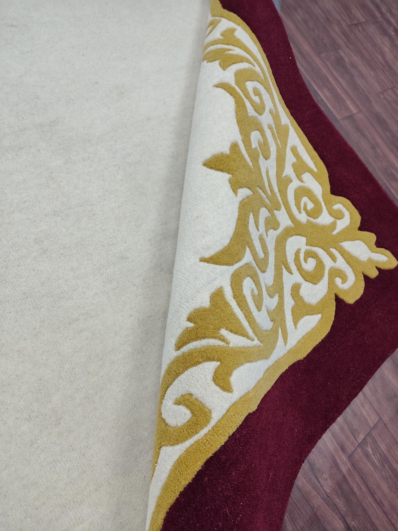K&ouml;nnte beinhalten: Eine aufgerollte Teppichfl&auml;che mit cremefarbenem Mittelteil und burgunderrotem Rand. Der Rand weist ein goldenes und wei&szlig;es Zierdesign auf. Der Teppich besteht aus einem weichen, pl&uuml;schigen Material.