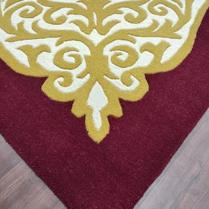 K&ouml;nnte beinhalten: Nahaufnahme eines Teils eines Teppichs mit bordeauxrotem Hintergrund und cremefarbenem Rand. Der Rand weist ein goldenes, verziertes Design auf. Der Teppich hat ein weiches, strukturiertes Aussehen.