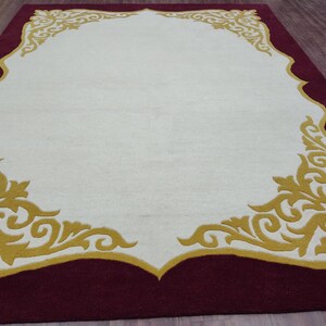 K&ouml;nnte beinhalten: Ein rechteckiger Teppich mit einem weinroten Rand und einer cremefarbenen Mitte. Die cremefarbene Mitte ist von einem goldenen, verzierten Design umrahmt. Das Design des Teppichs ist symmetrisch, wobei sich das goldene Muster in jeder Ecke und am oberen Rand wiederholt.