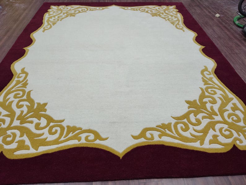 K&ouml;nnte beinhalten: Ein rechteckiger Teppich mit einem weinroten Rand und einer cremefarbenen Mitte. Die cremefarbene Mitte ist von einem goldenen, verzierten Design umrahmt. Das Design des Teppichs ist symmetrisch, wobei sich das goldene Muster in jeder Ecke und am oberen Rand wiederholt.