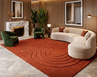 May include: A modern living room featuring a large, rust-colored rug with a wave pattern. A curved, cream-colored sofa with throw pillows sits near the rug. Two green armchairs and a marble fireplace with a gold accent are also visible.