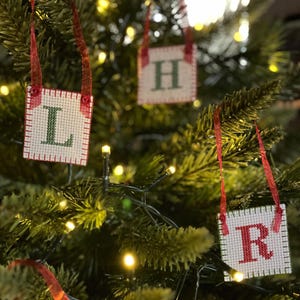 May include: Christmas tree ornaments with the letters "L", "H", "R", and "B" stitched on a square, off-white background. Each ornament has a red ribbon for hanging. The letters are green and the edges are red.