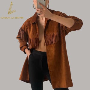 May include: A brown suede jacket with fringe detailing on the sleeves and chest. The jacket has a button-down front and a collar. The text "LONDON LUX LEATHER" is visible in the upper left corner. The jacket is paired with black pants.