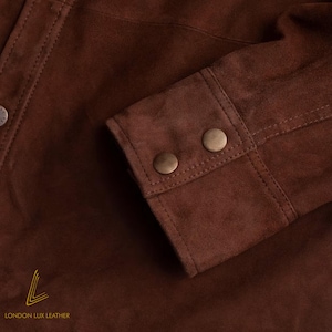 May include: Close-up of a brown suede jacket sleeve. The sleeve features two round, bronze-colored buttons and detailed stitching. The jacket is made of a soft, textured material. The logo "LONDON LUX LEATHER" is visible at the bottom.