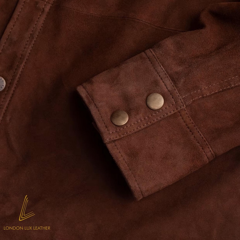 May include: Close-up of a brown suede jacket sleeve. The sleeve features two round, bronze-colored buttons and detailed stitching. The jacket is made of a soft, textured material. The logo "LONDON LUX LEATHER" is visible at the bottom.