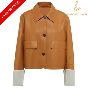 以下が含まれることがあります： クラシックな襟、2つのフロントポケット、黒いボタンが付いたタンレザージャケット。 ジャケットには白い袖口が付いています。 「FREE SHIPPING」という文字が左上に赤いバナーで表示されています。 London Lux Leatherのロゴが右上にあります。