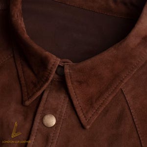 May include: Close-up of a brown suede jacket collar with a button closure. The jacket has a classic design with a pointed collar and visible stitching. The logo "LONDON LUX LEATHER" is visible at the bottom left.