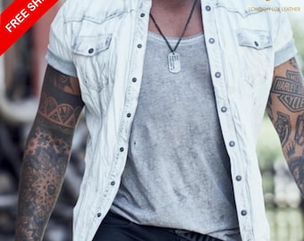 May include: A light-wash denim shirt with short sleeves, worn over a gray tank top. The shirt has button closures and two chest pockets. The image also shows a black necklace and tattoos on the arms.