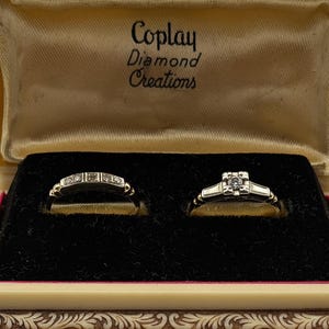 May include: Two silver-toned rings with small clear stones set in a cream-colored jewelry box. The interior lid features the text Coplay Diamond Creations. The rings are displayed on a black velvet insert within the box.