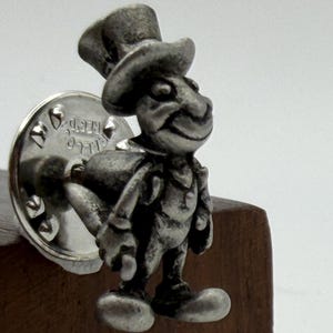 May include: A silver-toned metal lapel pin featuring a three-dimensional character wearing a tall top hat and a coat. The figure has a rounded nose and large feet, with a circular metal clutch backing visible behind the character's body.
