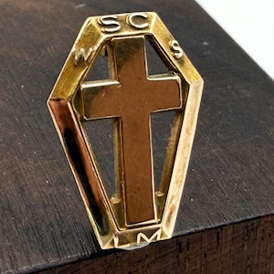 May include: A gold-toned metal pin shaped like a coffin with a central cross cutout. The letters W, S, C, and S are engraved along the top and side edges, while the letter M is engraved at the bottom point of the coffin-shaped frame.