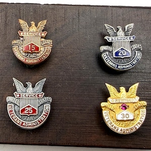 May include: Four United States Veterans Administration service pins featuring an eagle emblem, shield, and text indicating 15, 20, 25, and 30 years of service. The pins are finished in bronze, silver, blue, and gold tones with enamel accents.