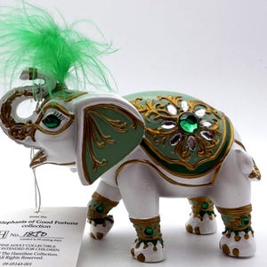 May include: A white and green elephant figurine from the Elephants of Good Fortune collection. The figurine has gold accents, green jewels, and a green feather plume. The tag reads "A FINE ADULT COLLECTIBLE NOT INTENDED FOR CHILDREN".