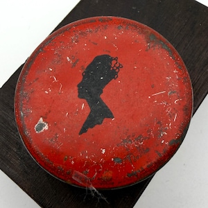 Op de afbeelding: Een ronde, rode blikken doos met een zwart silhouet van een vrouwenprofiel. De doos heeft een verweerde uitstraling en ligt op een donker houten blok. Het silhouet toont een gedetailleerd kapsel.