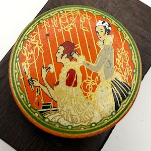 May include: A vintage, round compact case with an orange background and a decorative illustration. The illustration depicts two figures in period clothing, one applying makeup. The case has a gold and green border.