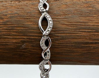 May include: A silver-toned link bracelet featuring a repeating pattern of infinity-shaped loops. Each link is accented with small, clear, sparkling stones set along the metal surface, creating a continuous, articulated design throughout the piece.