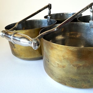 May include: Four antique brass measuring cups with metal handles. The cups have a tarnished, aged appearance, with a silver handle on the front-most cup. The handles are attached to the cups with a metal bracket.