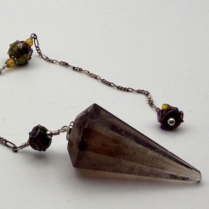 May include: A pointed, faceted smoky quartz crystal pendulum attached to a silver-toned metal chain. The chain features three decorative beads with multicolored floral patterns and small yellow accents, connected by thin wire links.
