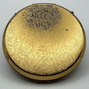 May include: A round gold-toned metal compact mirror featuring an intricate, engraved paisley pattern across the entire top surface. The design consists of swirling, detailed motifs that cover the metallic finish, creating a textured appearance.