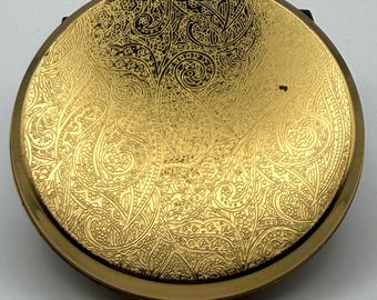 May include: A round gold-toned metal compact mirror featuring an intricate, engraved paisley pattern across the entire top surface. The design consists of swirling, detailed motifs that cover the metallic finish, creating a textured appearance.