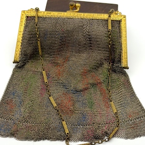 Op de afbeelding: Een antieke metalen mesh portemonnee met een goudkleurig frame en kettingriem. De portemonnee heeft een decoratieve sluiting en een houten bovenkant. De mesh heeft een kleurrijk bloemenpatroon dat zichtbaar is door het metaal.